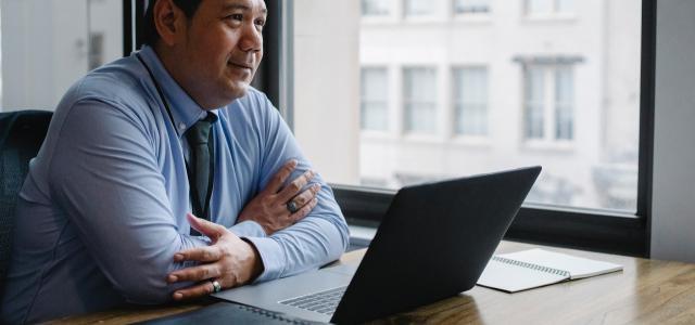 Professional-looking man sitting in an office. There is a laptop and a stack of files in front of him.